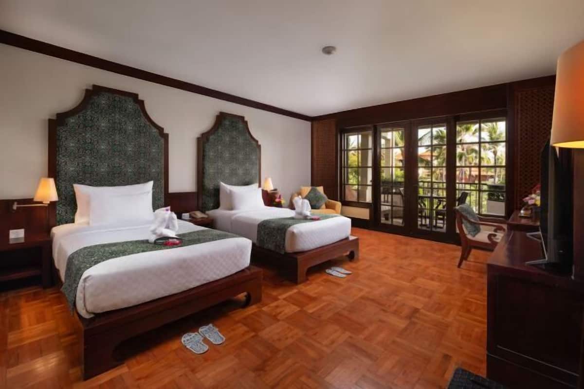 A hotel room with two beds, wooden flooring, large windows with a view, and a TV on a wooden cabinet.