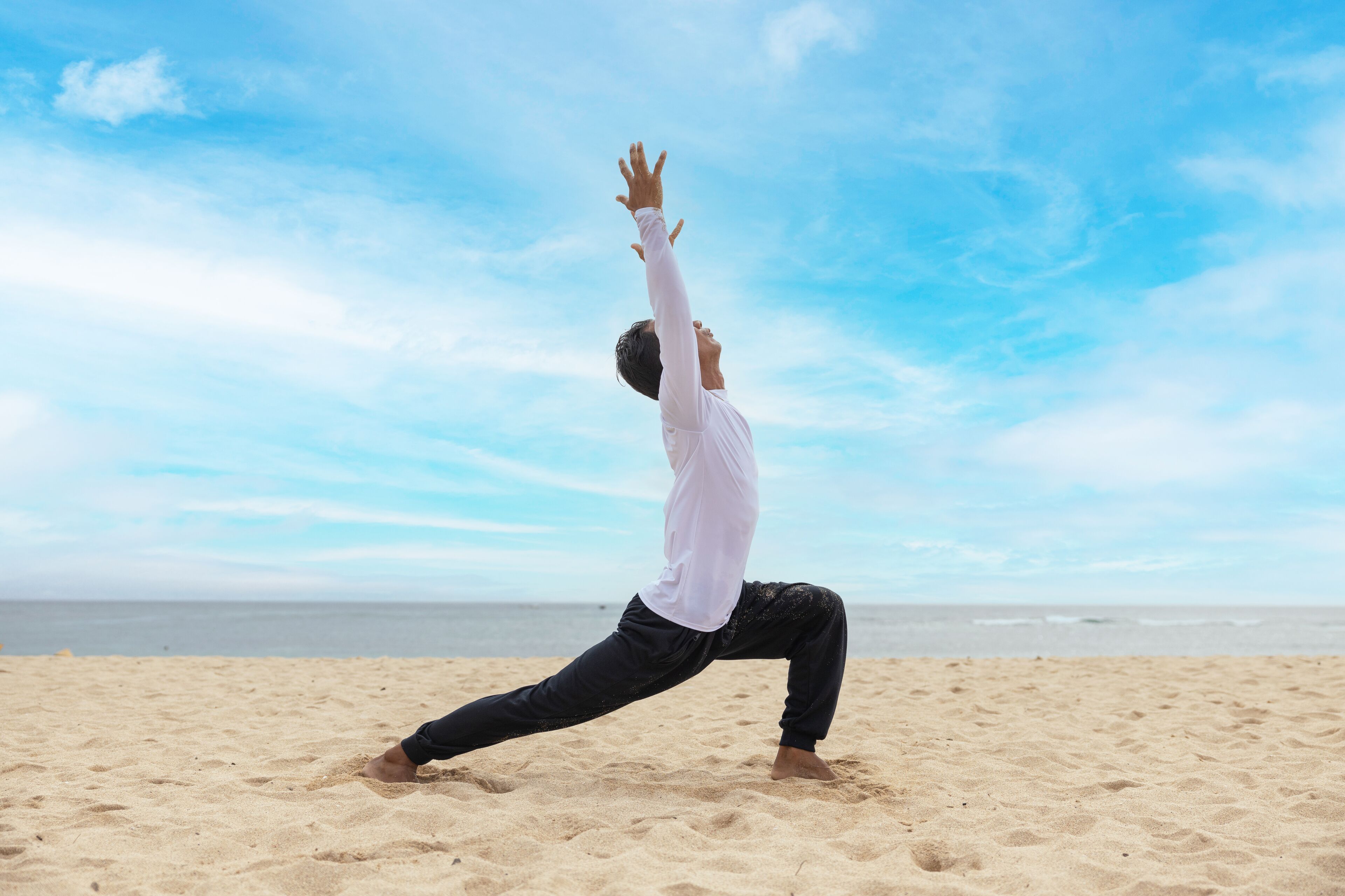 En person udfører en yogastilling på en sandstrand med oceanen og en klar himmel i baggrunden.
