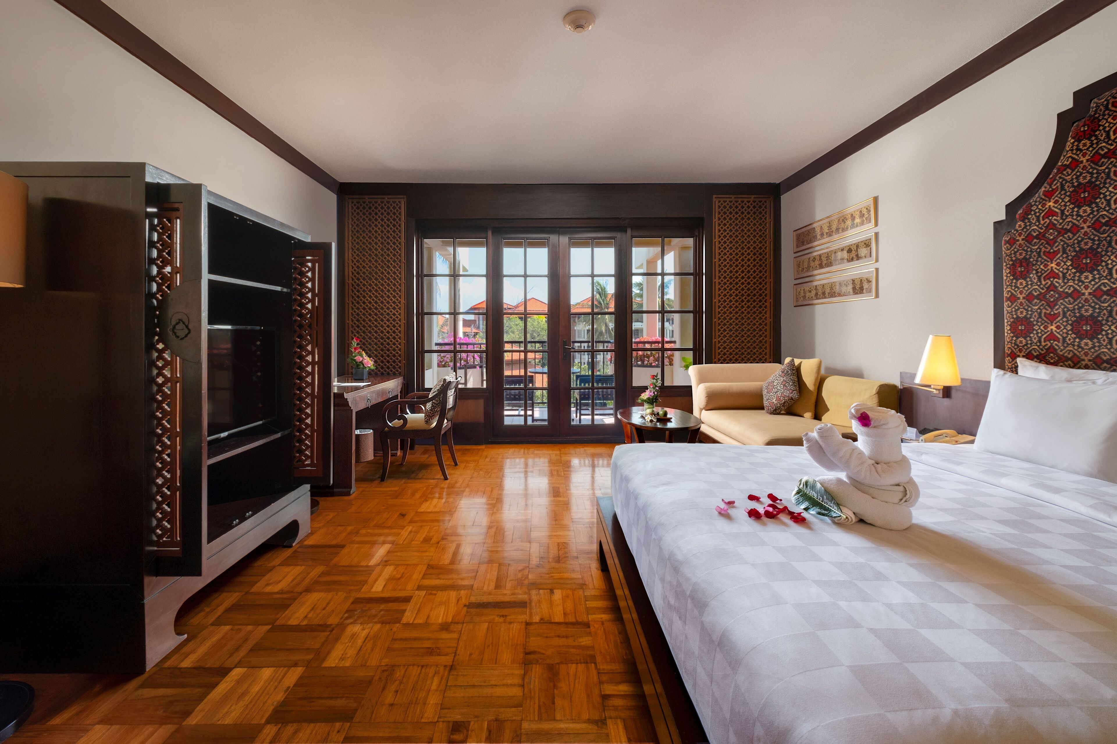 A hotel room with a large bed, a desk, a TV, and a balcony with a view of buildings.