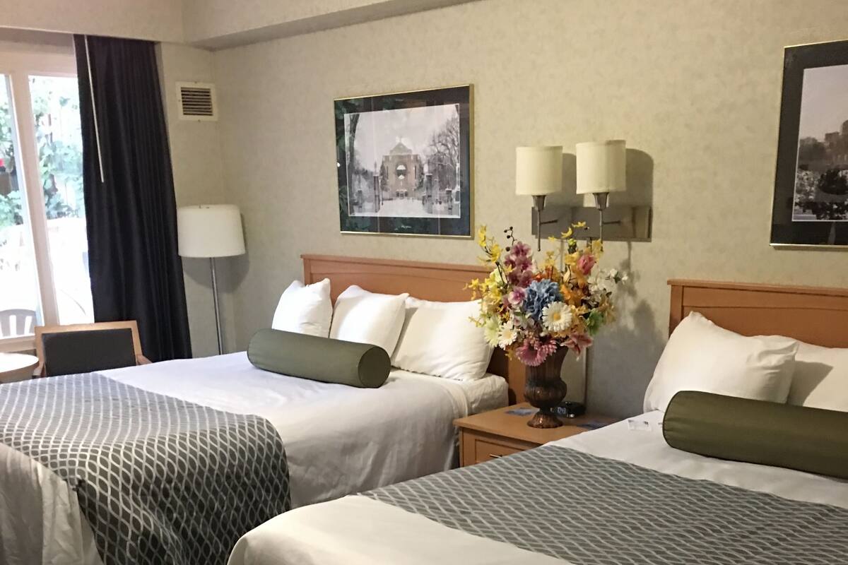 A hotel room with two beds, a small table, a chair, a desk, and a desk lamp. There is a window with curtains, a picture on the wall, and a vase with flowers on the desk.