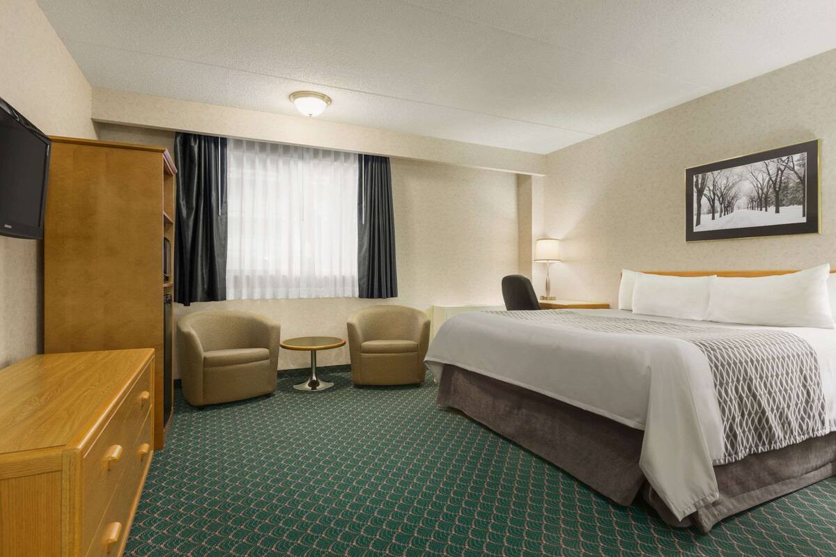 A hotel room with a bed, two armchairs, a small table, a TV, and a painting on the wall.