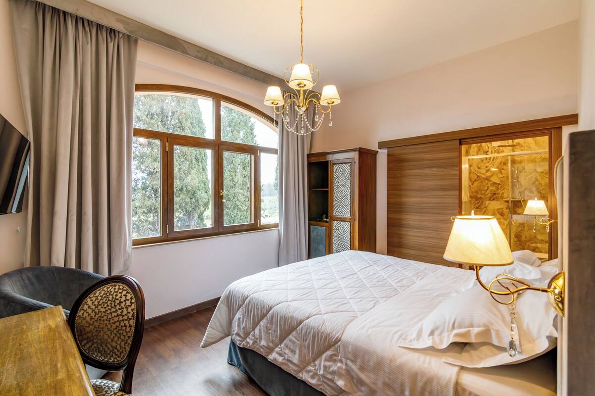 A bedroom with a large bed, a chair, a table, a chandelier, and a window with curtains.