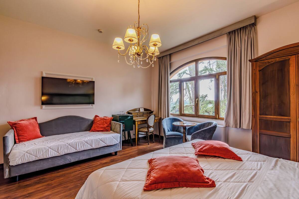 Ein Hotelzimmer mit einem großen Bett, einem Sofa, einem Schreibtisch und einem Sessel. Ein Fenster mit Vorhängen und einer Deckenleuchte.