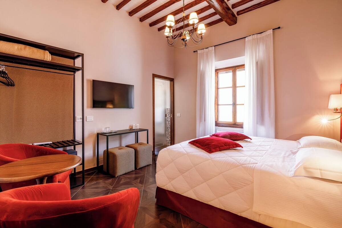 A hotel room with a large bed, a wooden table, red chairs, a TV, and a chandelier.