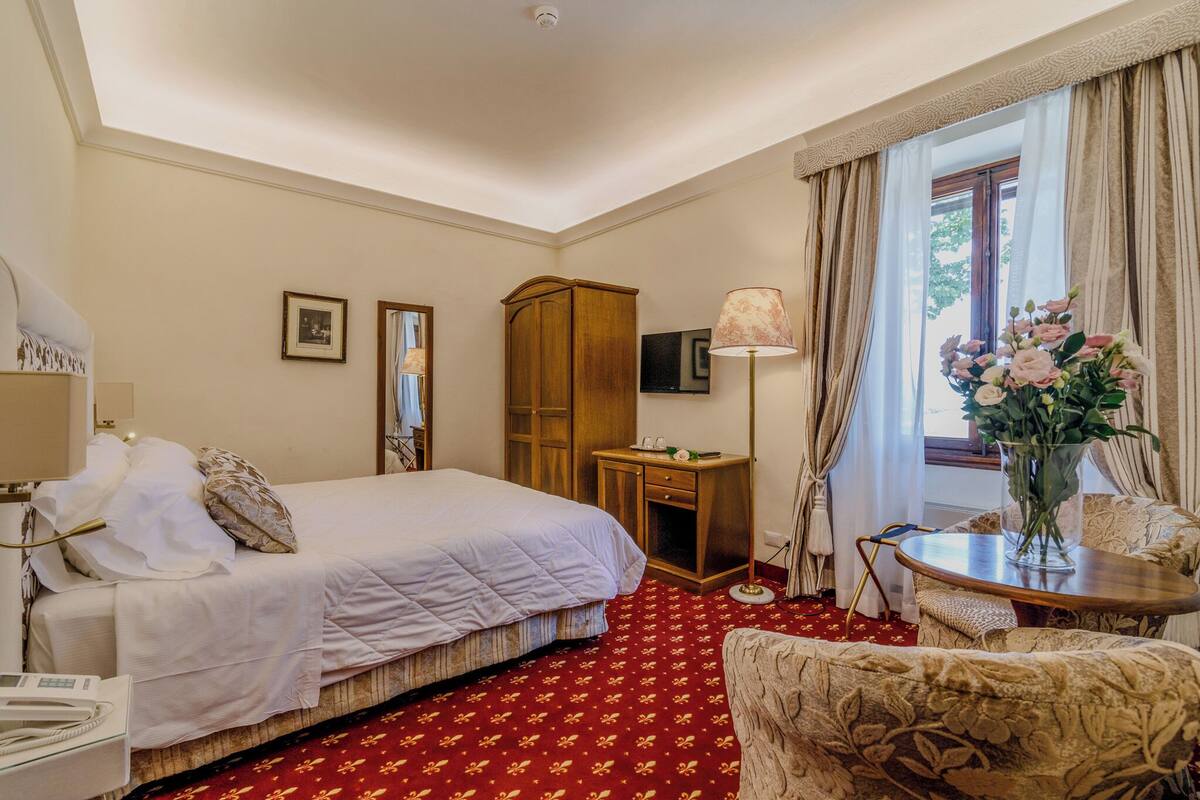 Ein Hotelzimmer mit Bett, Schreibtisch, Stuhl, Lampe, Kleiderschrank und einem Fenster mit Vorhängen.