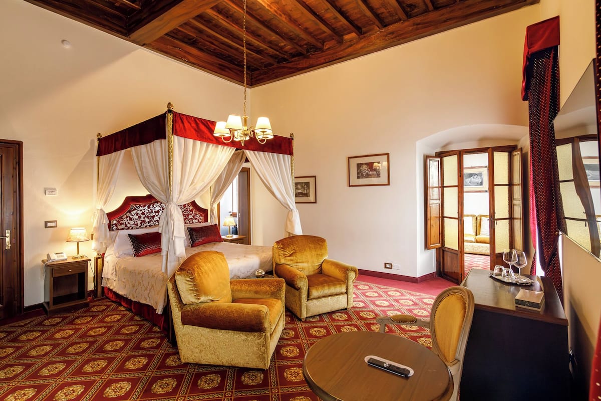 A room with a four-poster bed, two armchairs, a round table, and a view of the outside through a window.