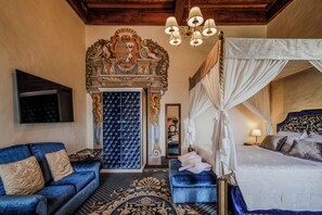 A luxurious bedroom with a large bed, a blue sofa, a decorative fireplace, and a chandelier.