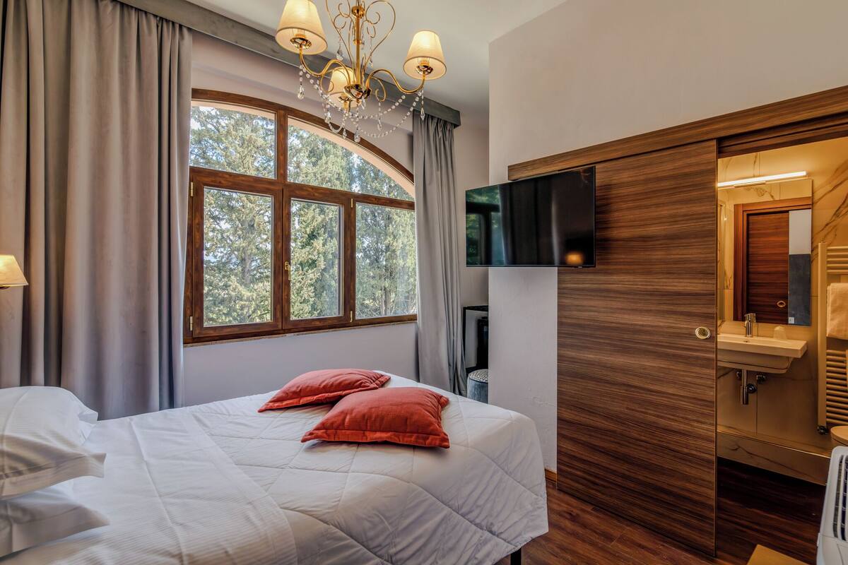 Ein Schlafzimmer mit großem Fenster, einem Bett mit weißer und roter Bettwäsche, einer Kristallleuchtreibe, einem an der Wand montierten Fernseher und einem Holzschrank.