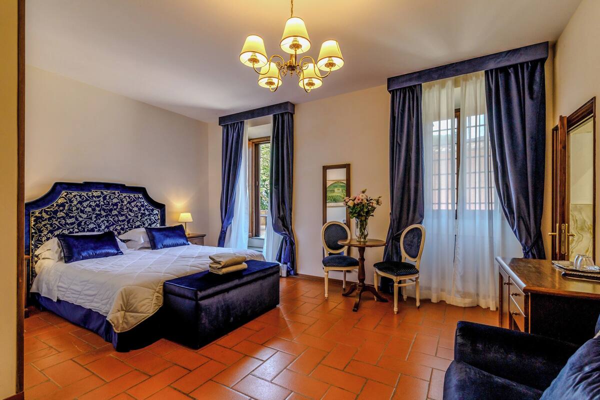 A hotel room with a large bed, a desk, two chairs, a chandelier, and curtains.