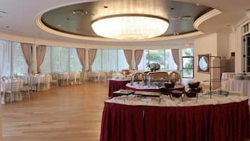 A banquet hall with round tables covered in red tablecloths, a large chandelier, and a buffet setup.