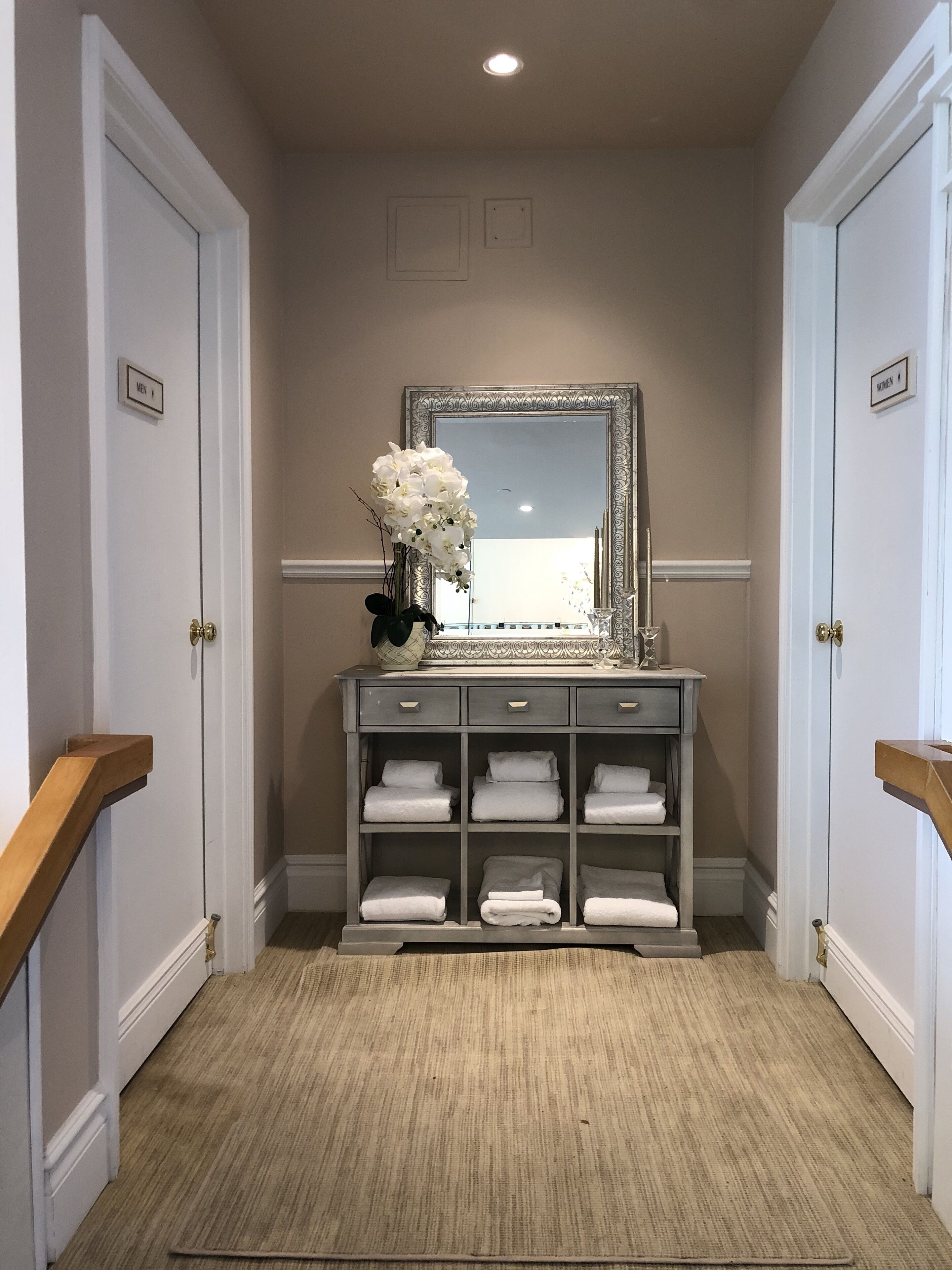 A hallway with a vanity table, a mirror, and a vase of white flowers. There are two doors labeled "MEN" and "WOMEN".