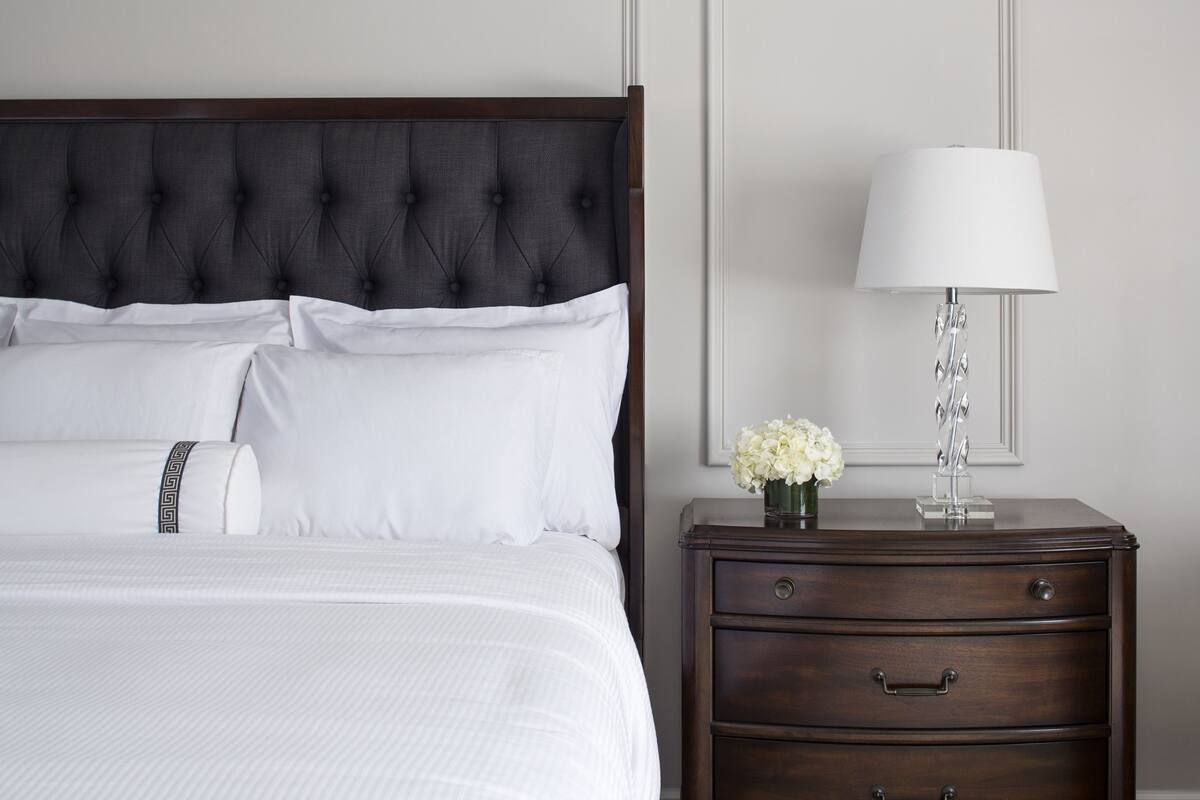 Una cama bien hecha con cabecera oscura, ropa de cama blanca y una mesita de noche con una lámpara y un jarrón de flores.