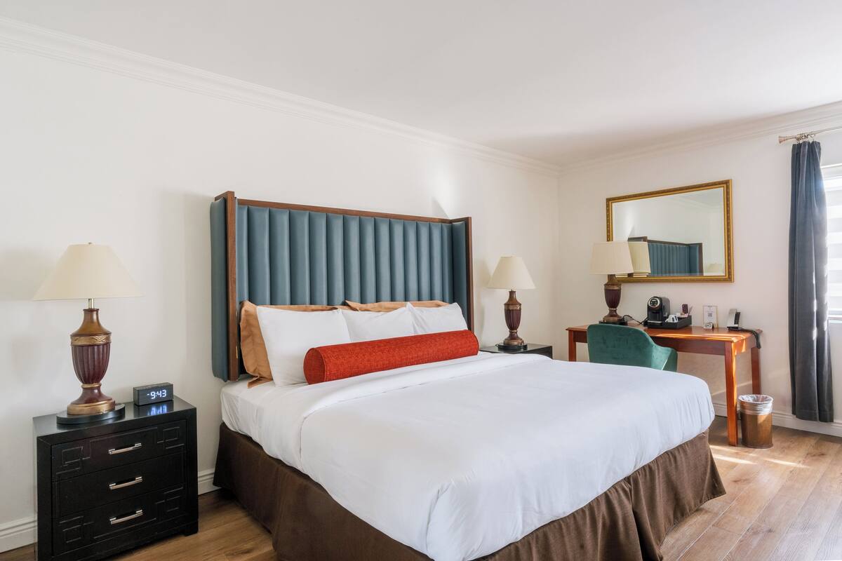Habitación de hotel con una cama grande, un escritorio, una silla, una mesita de noche, una lámpara, un espejo y una ventana con cortinas.