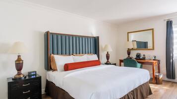 A hotel room with a large bed, a desk, a chair, a nightstand, a lamp, a mirror, and a window with curtains.