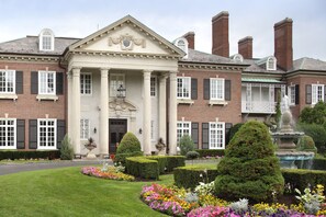 A large, stately building with a symmetrical facade, featuring a central entrance with columns, a decorative pediment, and a fountain in the garden. - The Mansion at Glen Cove (Glen Cove)