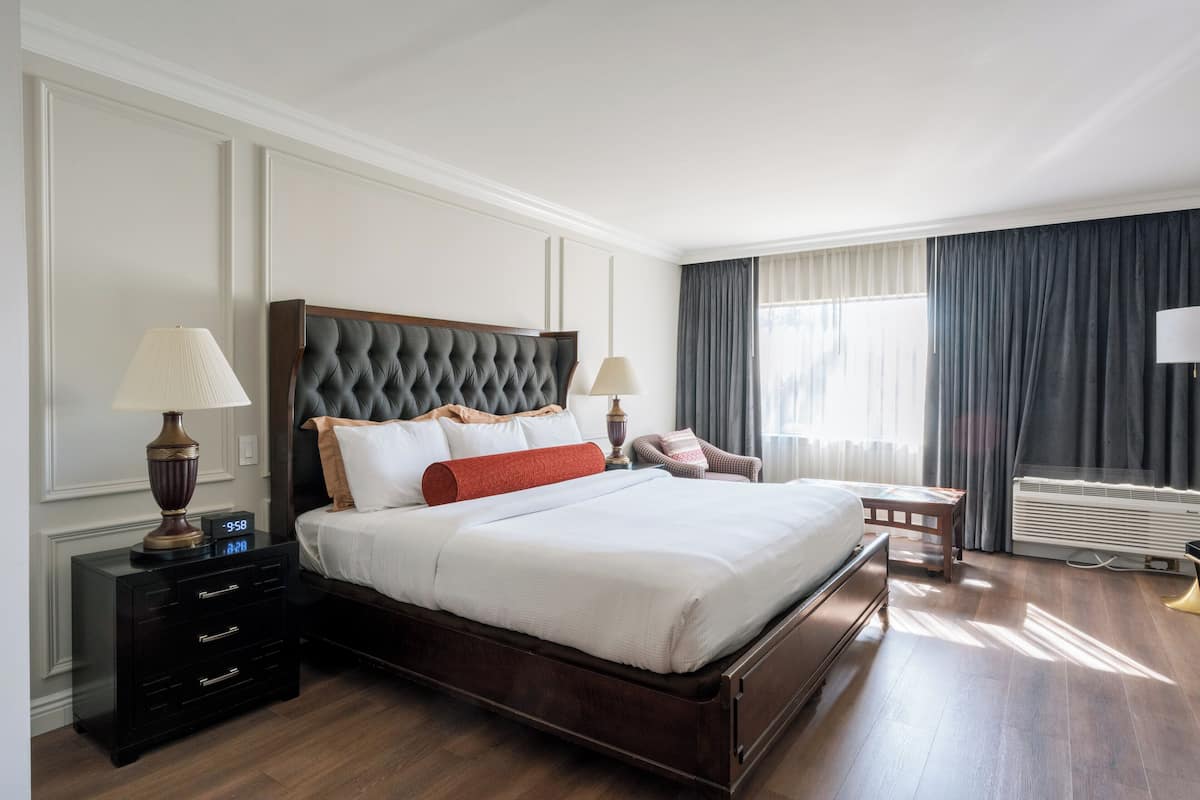 Un dormitorio ordenado con una cama grande, mesitas de noche, una silla y una ventana con cortinas.