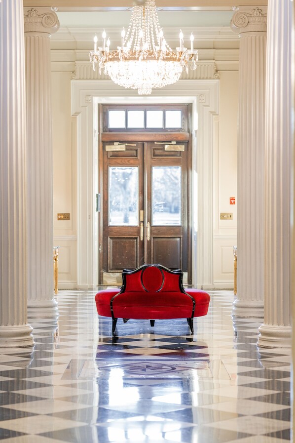 A grand hallway with a red upholstered bench, a large chandelier, and a wooden door with glass panels.