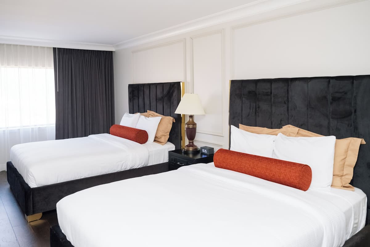 Dos camas dobles con sábanas blancas y almohadones rojos, una mesita de noche con lámpara y una ventana con cortinas traslúcidas.