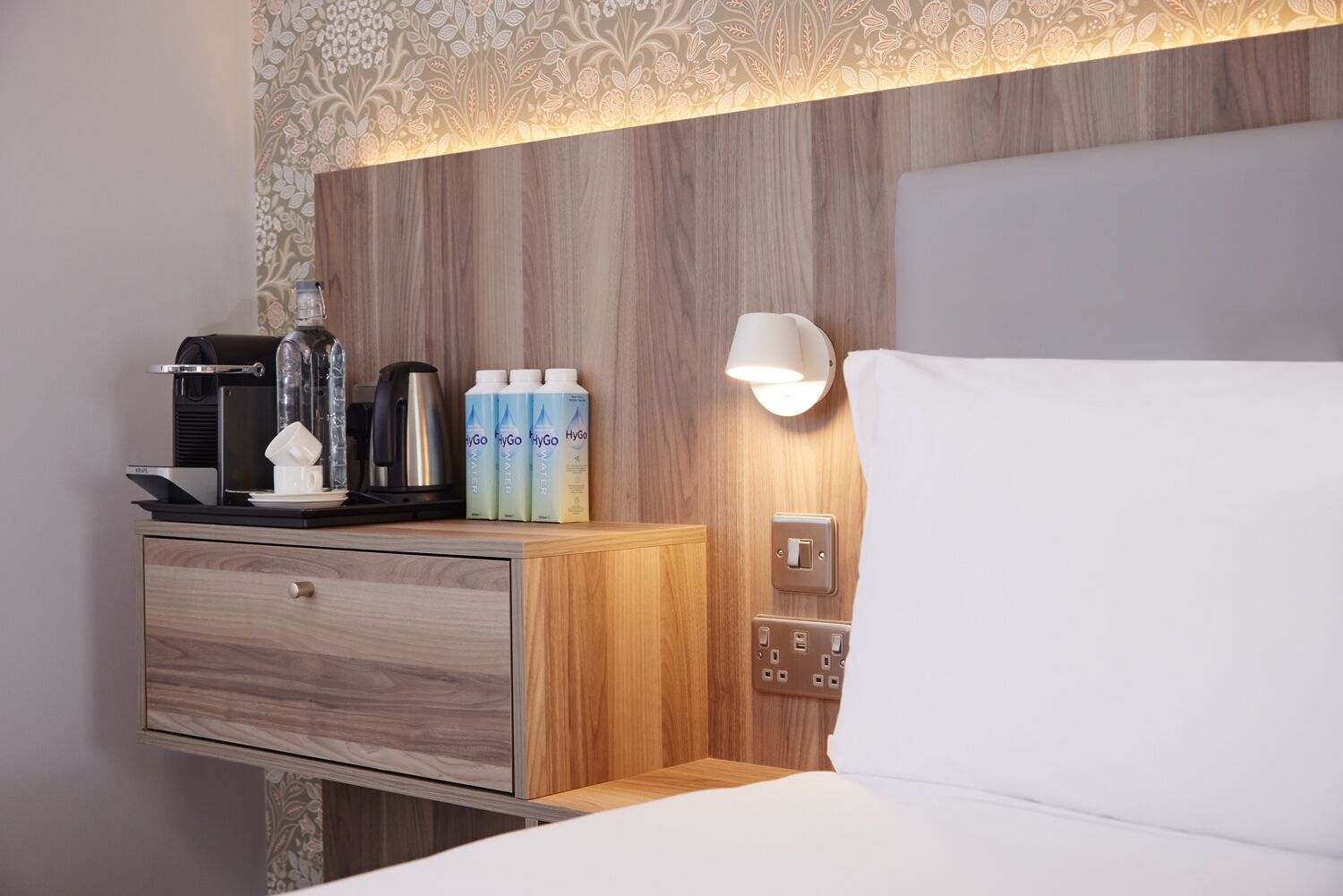 Une chambre d’hôtel avec une tête de lit en bois, une table de chevet avec une machine à café, une lampe et des bouteilles d’eau.