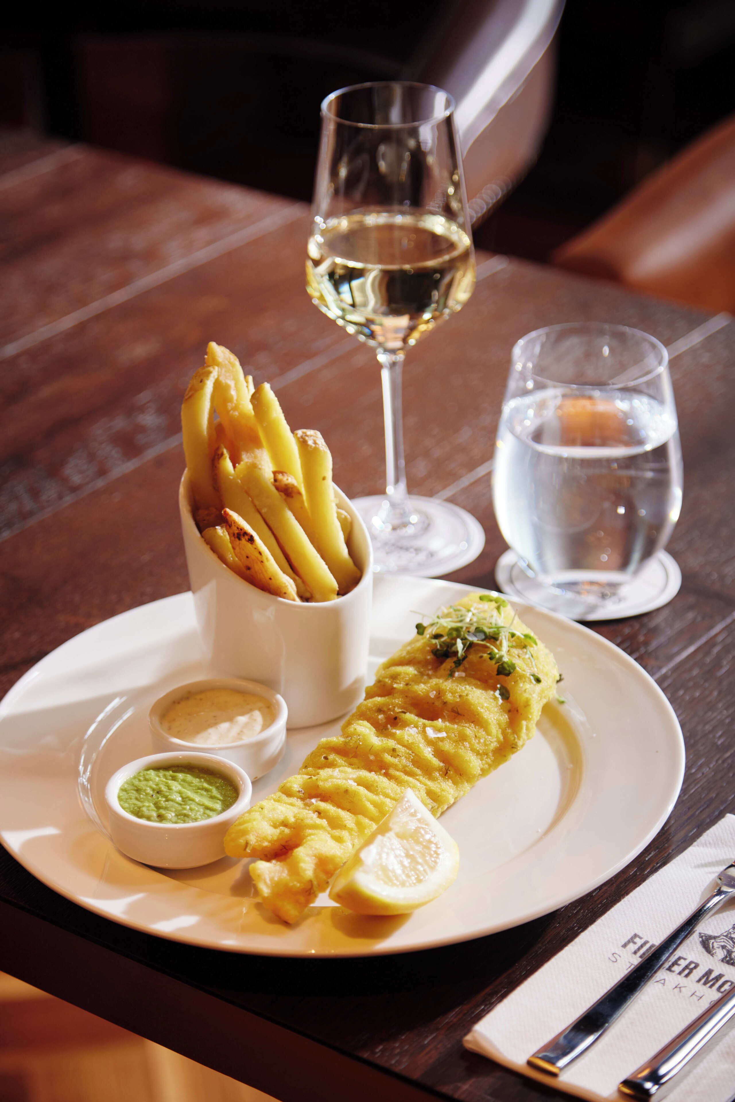 A plate with a fish fillet, fries, and two small dipping sauces, accompanied by a glass of white wine and a glass of water.