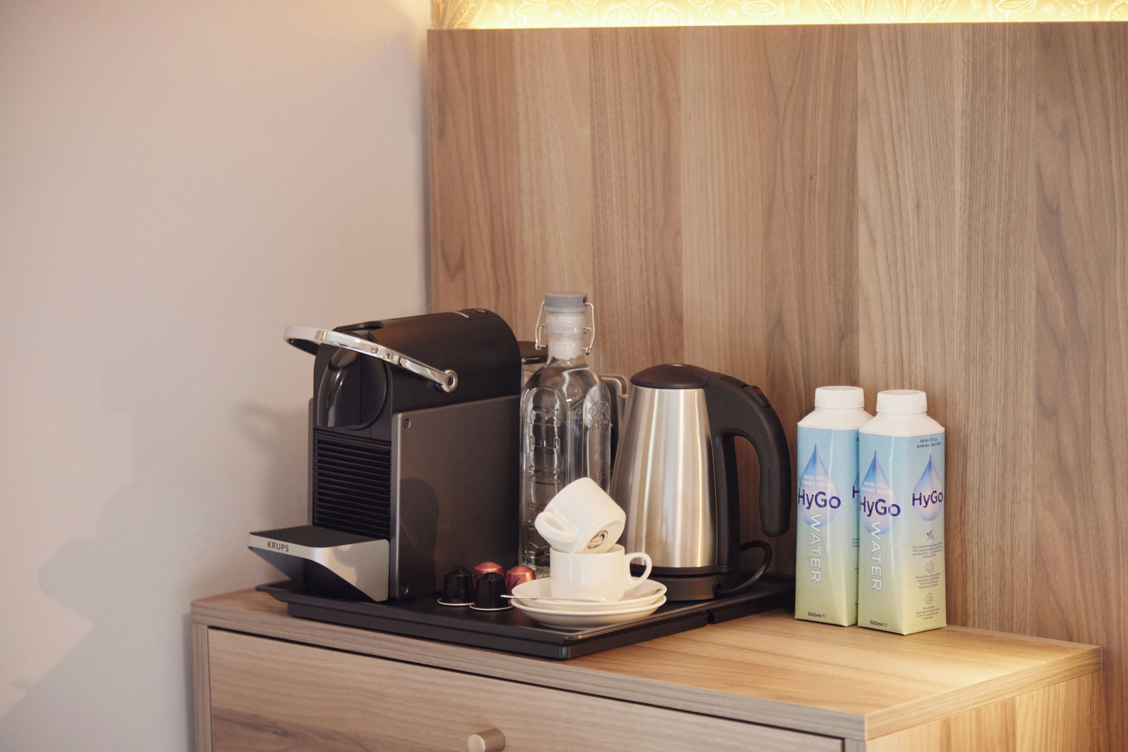 Angolo caffè con una macchina da caffè Krups, una bottiglia d'acqua, una caraffa di vetro e due bottiglie d'acqua HyGo su un tavolo di legno.