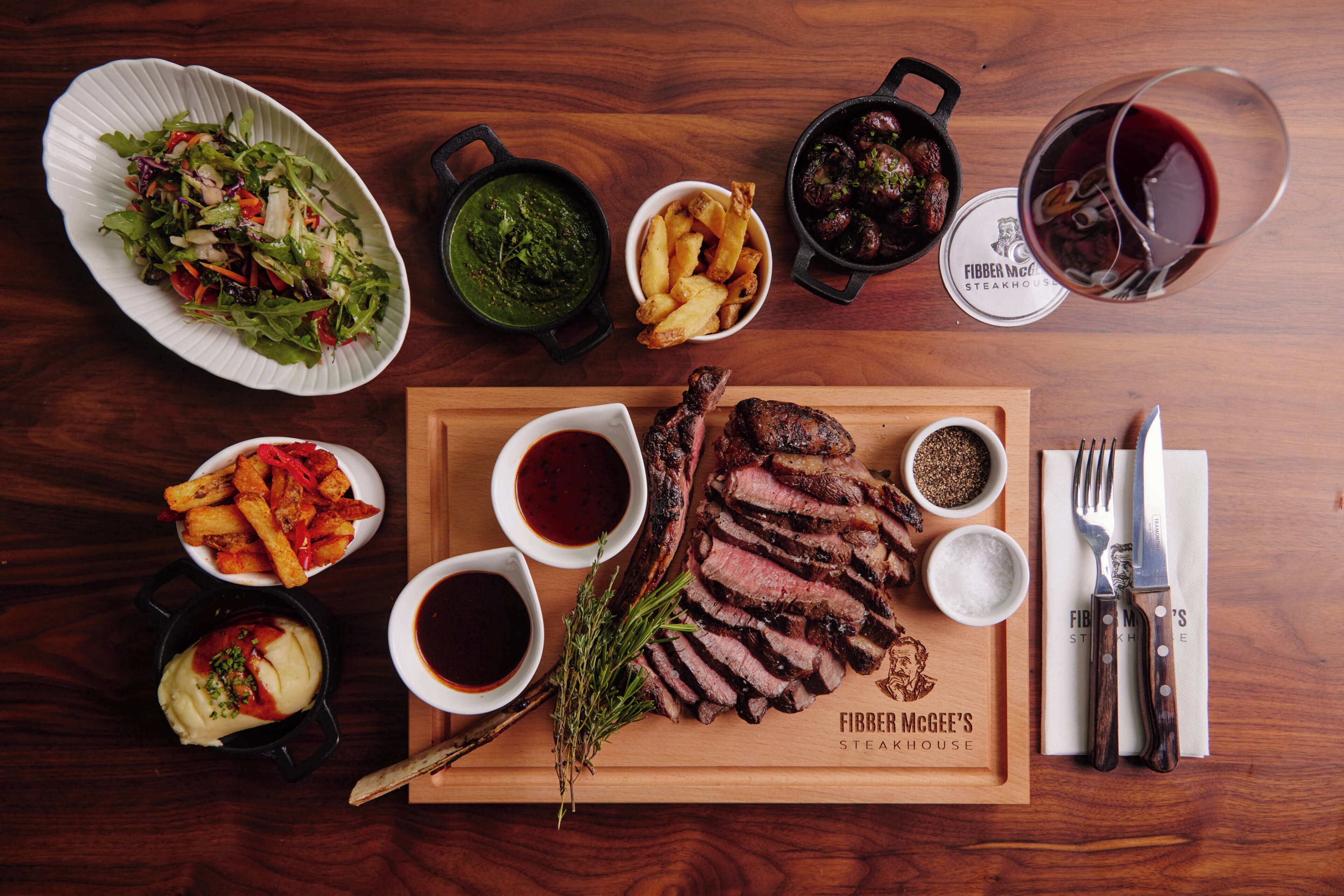 Una mesa de madera con diversos platos, incluyendo bistec, papas fritas, ensalada y salsas.