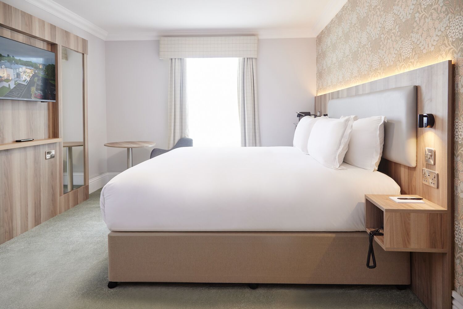 Habitación de hotel con una cama grande, un televisor montado en la pared, una mesita y una ventana con cortinas.