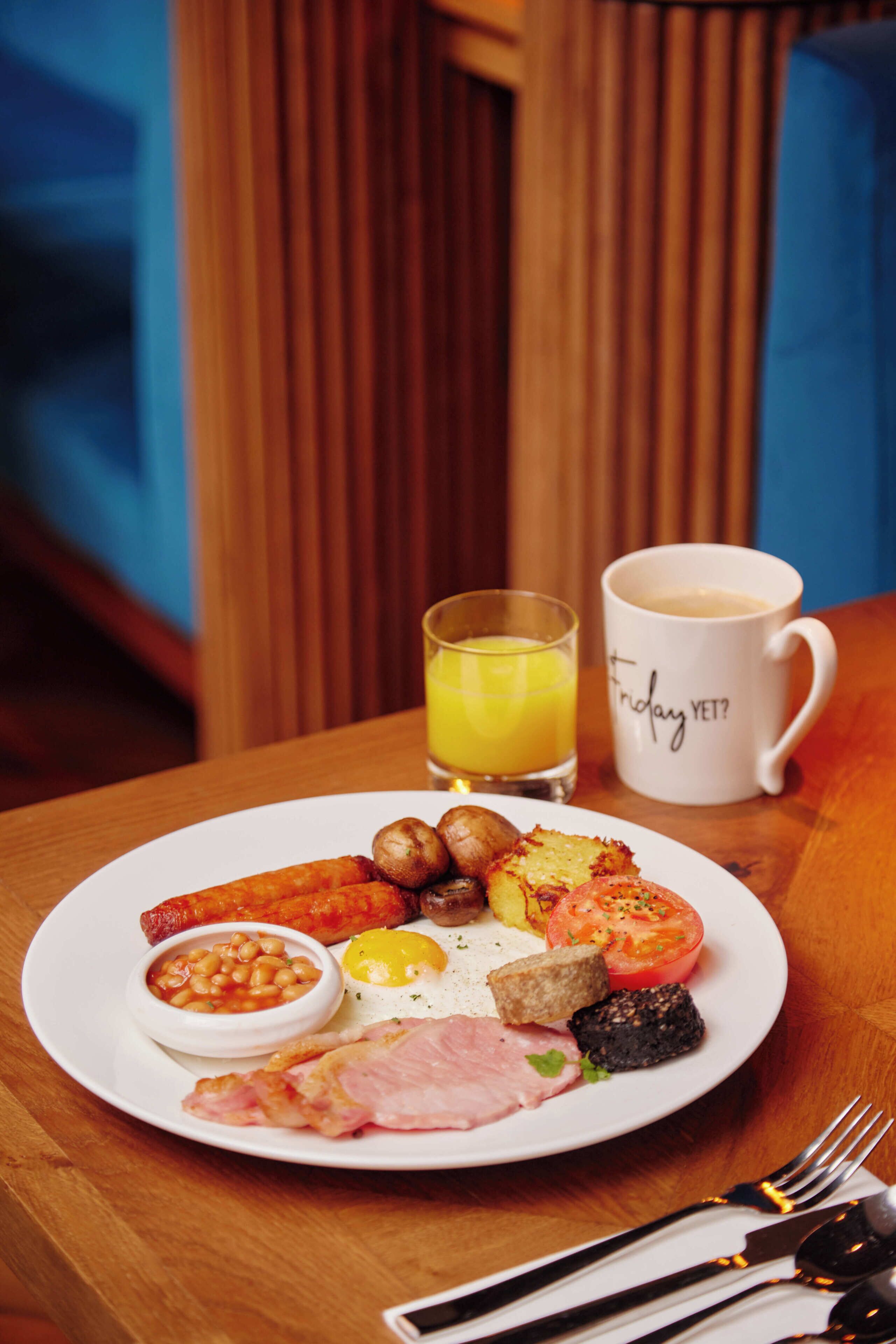 Un piatto per la colazione con uova, pancetta, salsicce, funghi e fagioli, accompagnato da un bicchiere di succo d'arancia e una tazza di caffè.