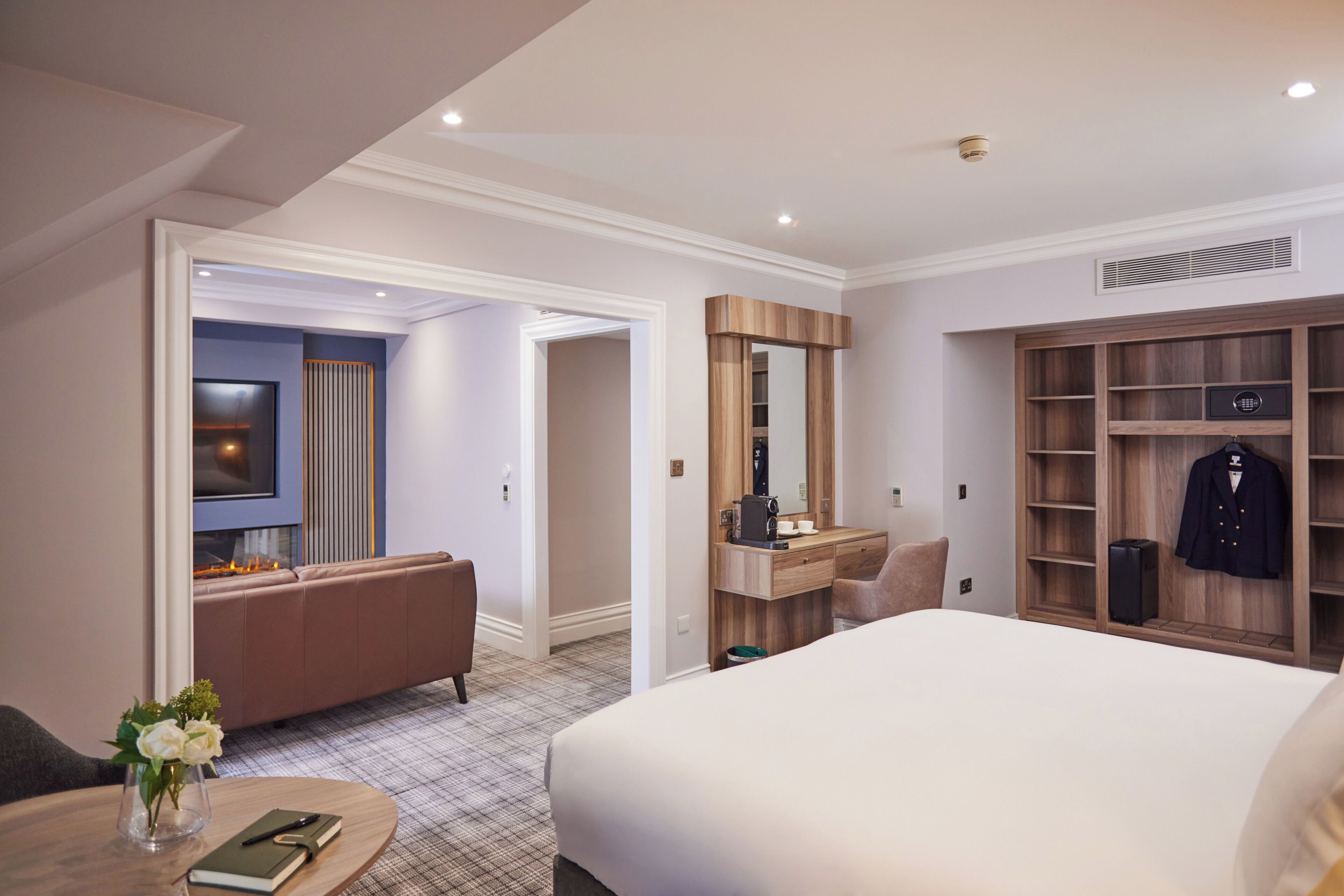 A hotel room with a bed, a desk, a mirror, a wardrobe, a seating area, and a small table with a vase.