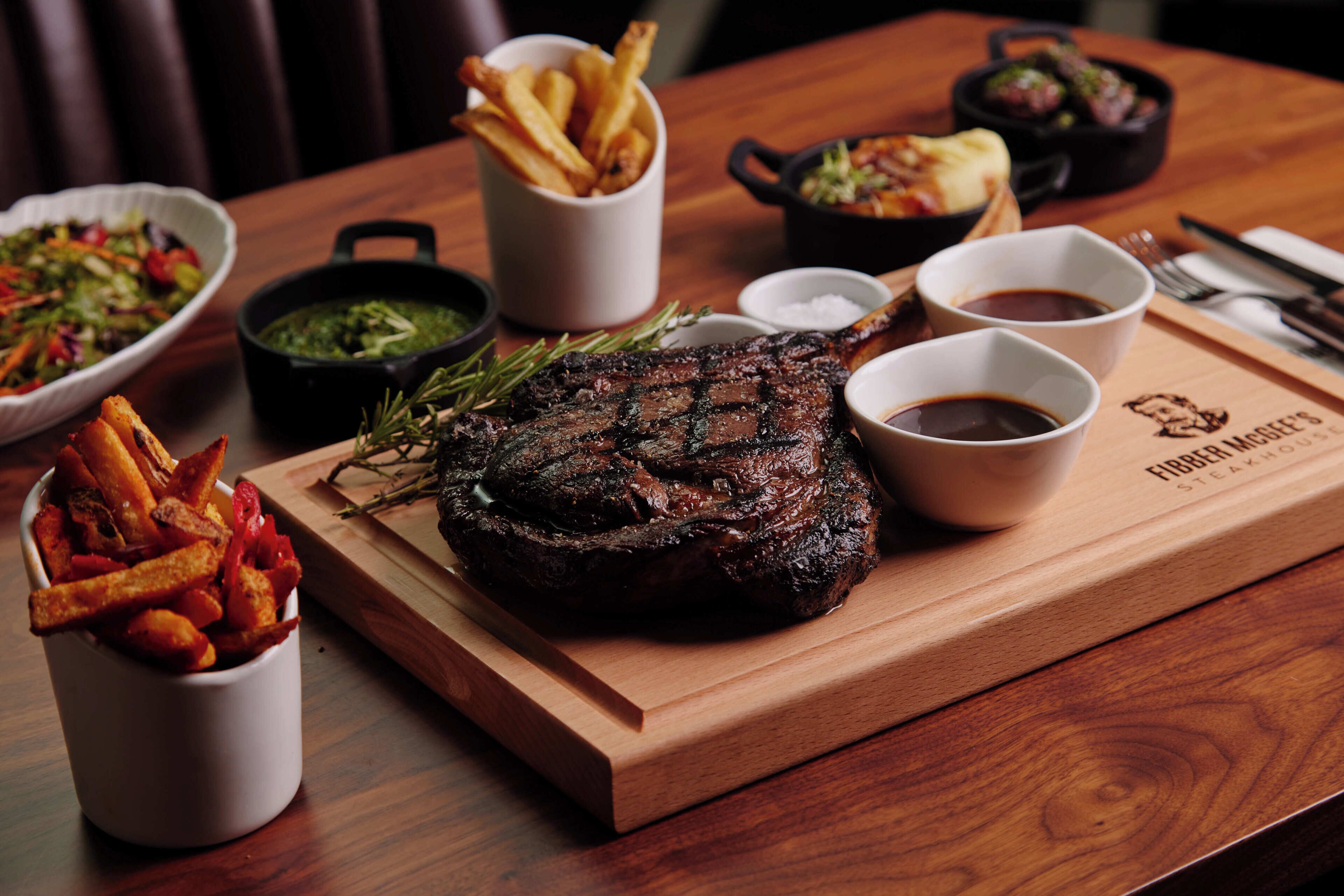 A wooden table with a steak, fries, and various side dishes.