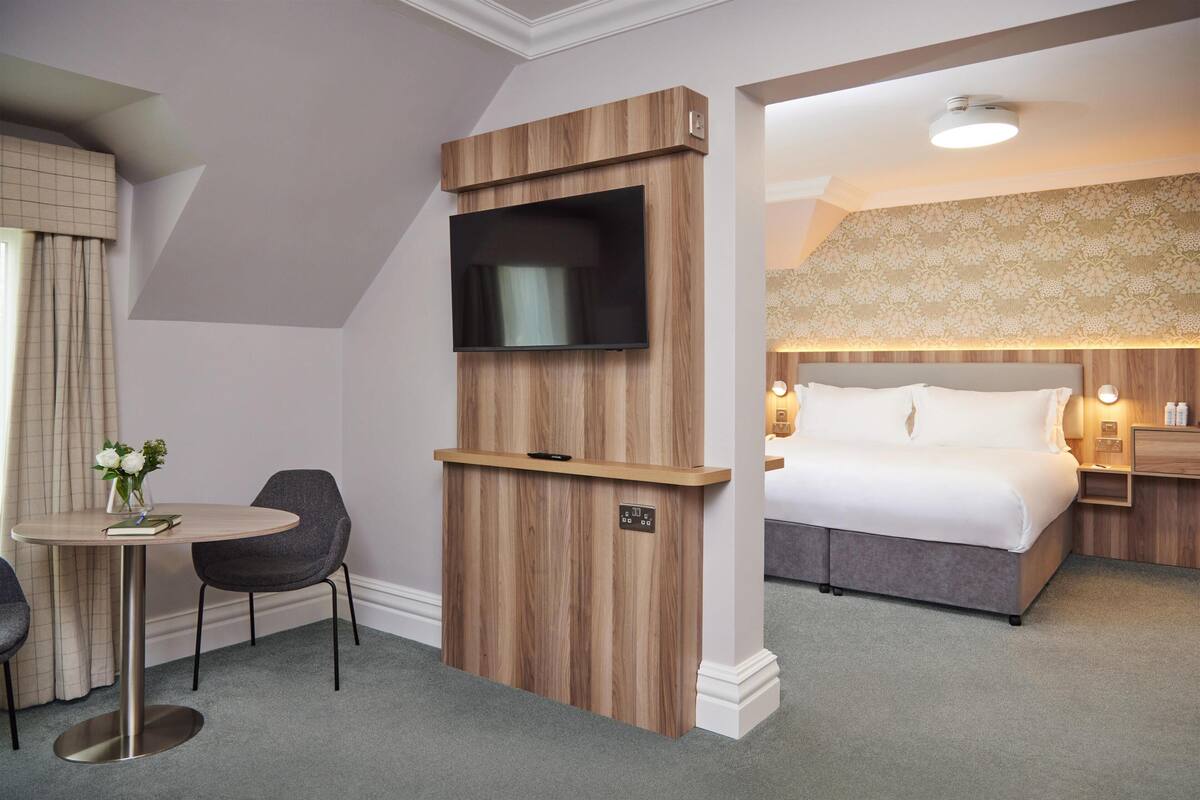 Una habitación de hotel con una cama, un televisor montado en un panel de madera, una mesita con una silla y una mesita de noche con una lámpara.