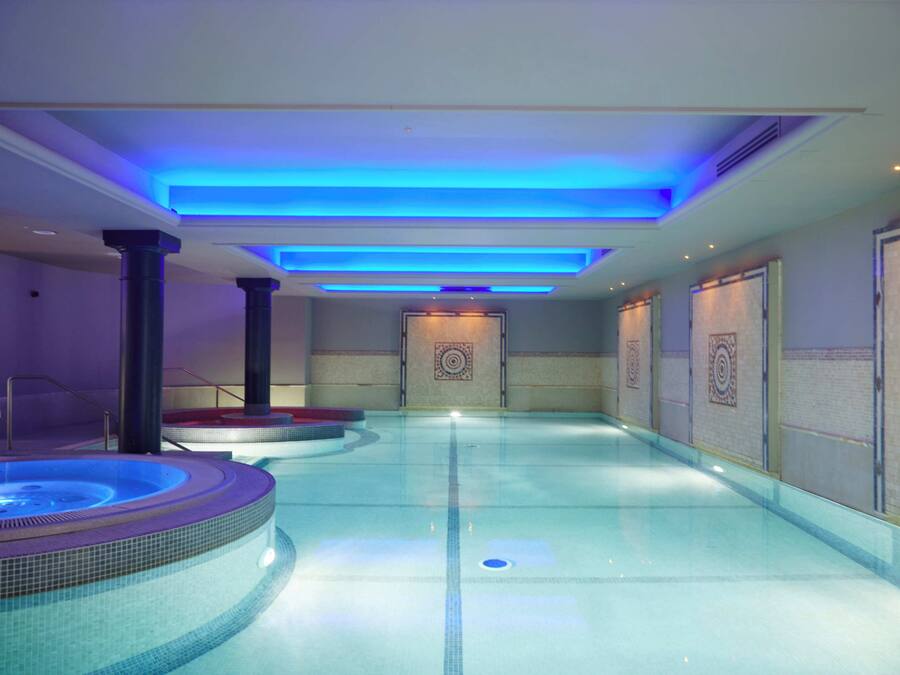 A modern indoor swimming pool with a hot tub, blue lighting, and a tiled floor.