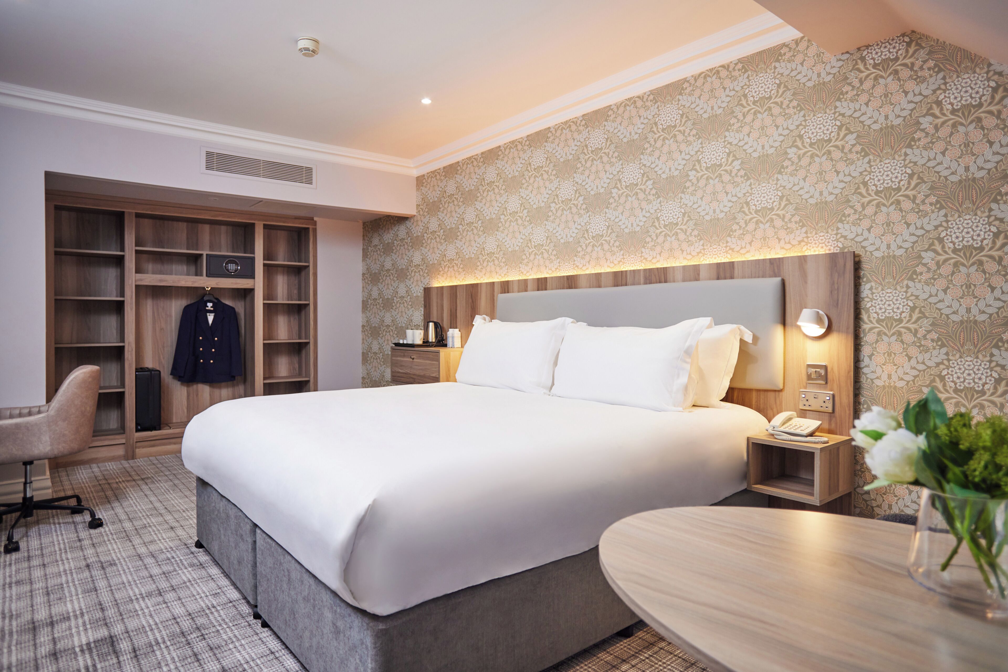 A hotel room with a large bed, a desk, a chair, a wardrobe, and a nightstand with a phone.