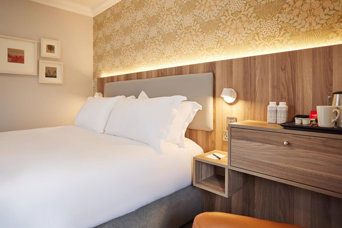 Habitación de hotel con una cama grande, cabecera de madera, mesita de noche con lámpara, cajón y estante en la pared.