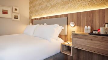 Ein Hotelzimmer mit einem großen Bett, einem hölzernen Kopfteil, einem Nachttisch mit Lampe, einer Schublade und einem Wandregal.