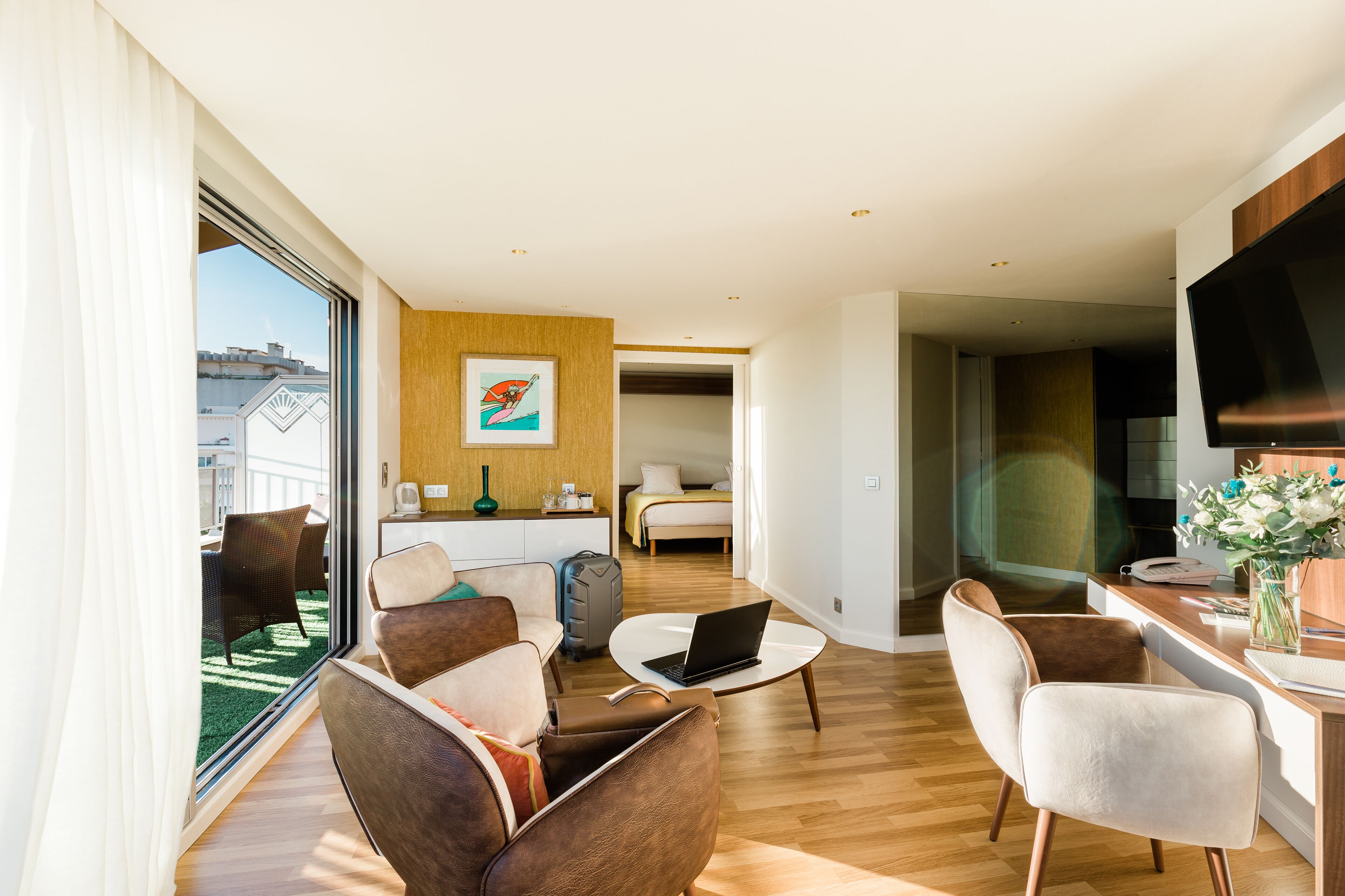 Ein modernes Hotelzimmer mit Balkon, Essbereich, Wohnzimmer mit Fernseher und Schlafzimmer mit Bett und Schreibtisch.