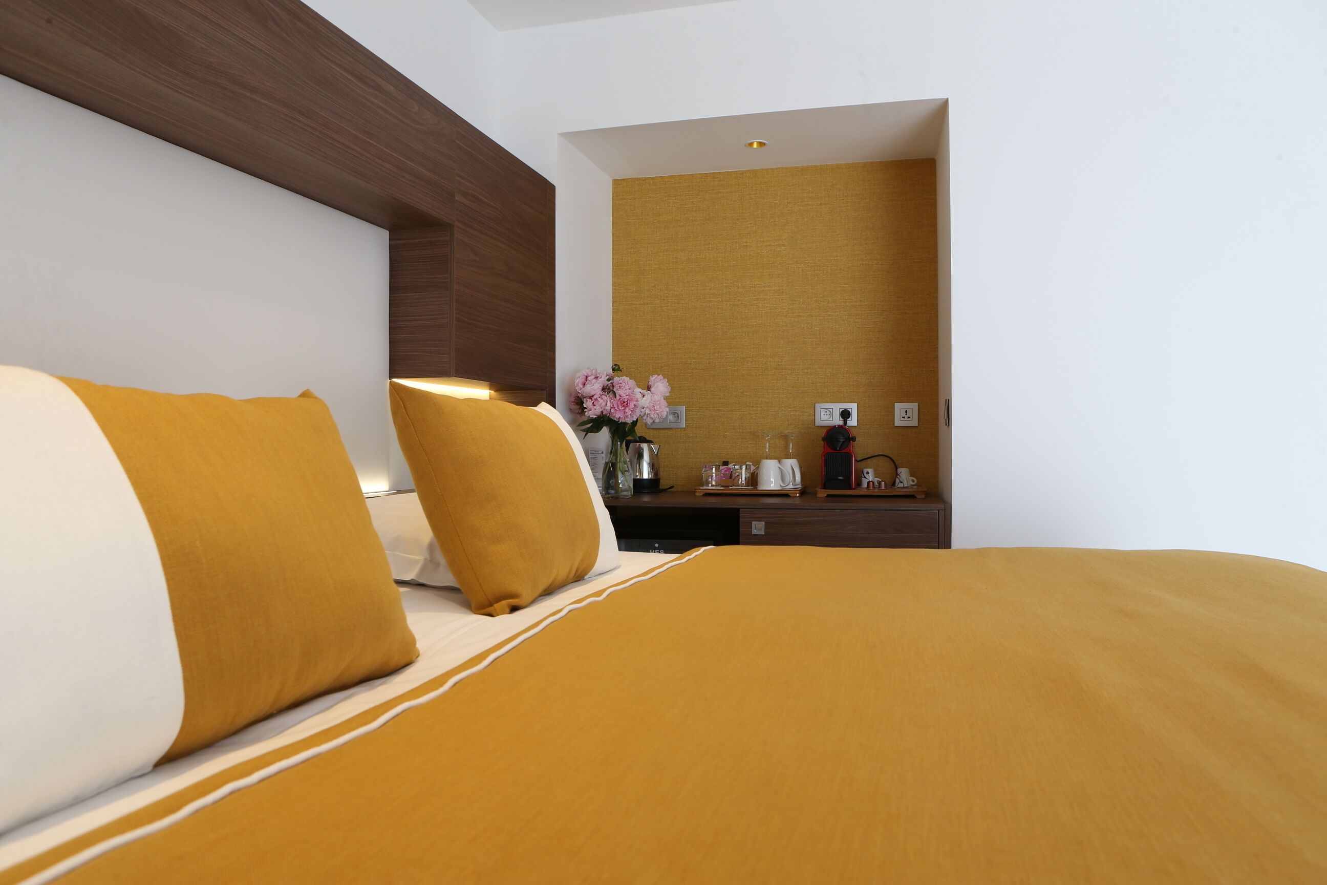 Une chambre d’hôtel avec un grand lit, une tête de lit en bois, un petit bureau agrémenté d’un vase de fleurs et une prise électrique fixée au mur.
