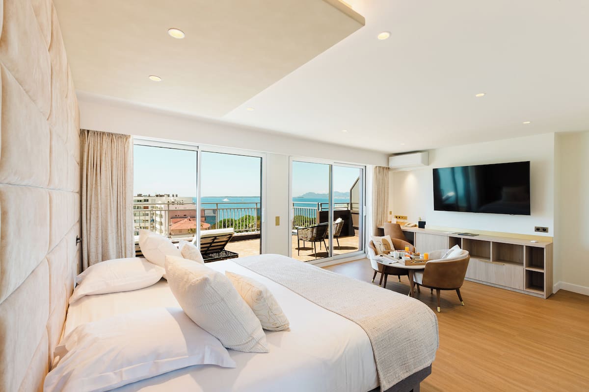 A modern hotel room with a large bed, a TV, a balcony with a view of the sea, and a dining area.