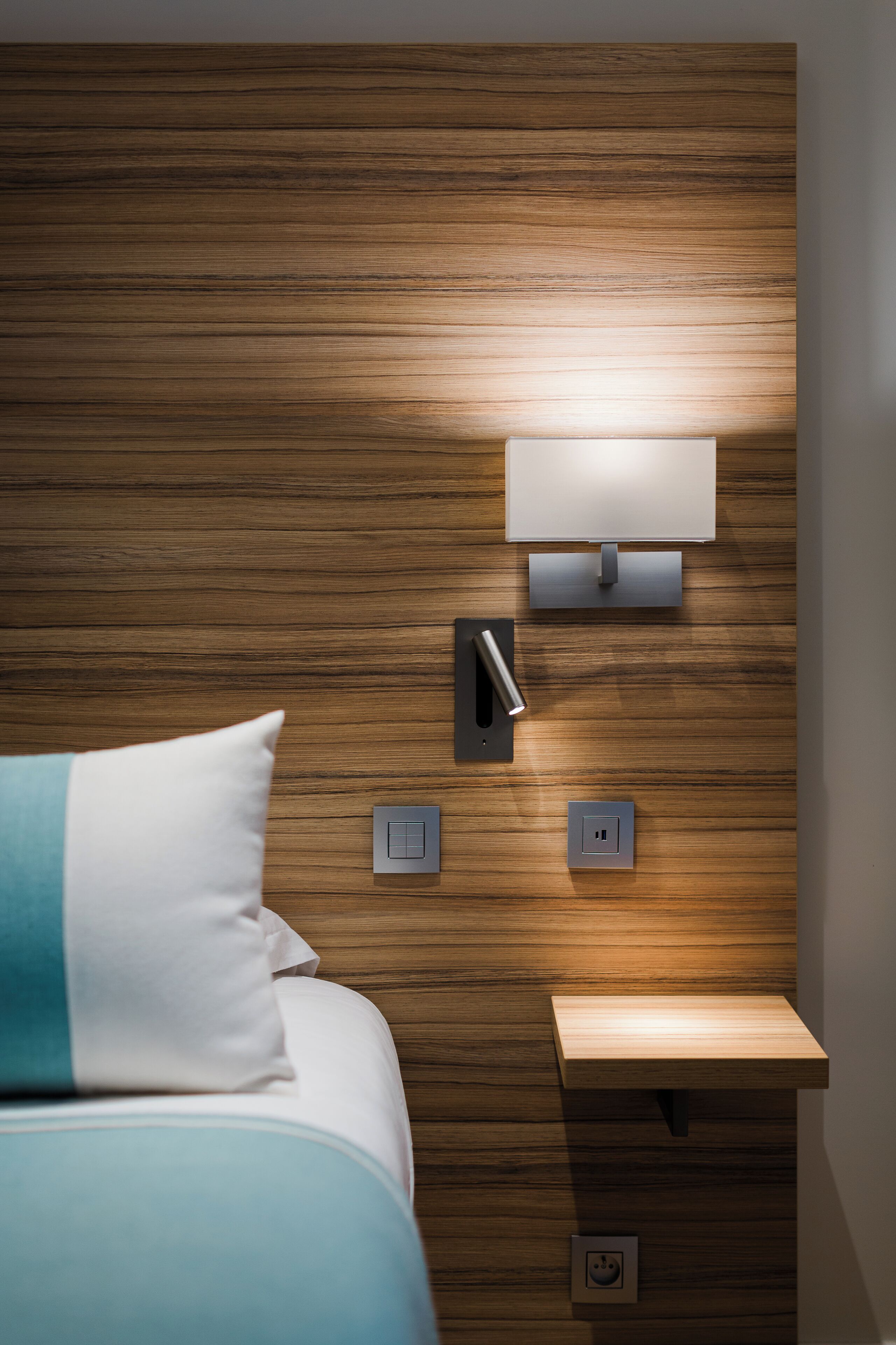 Une chambre d’hôtel moderne avec une tête de lit en bois, une table de chevet avec une lampe et une prise de courant.