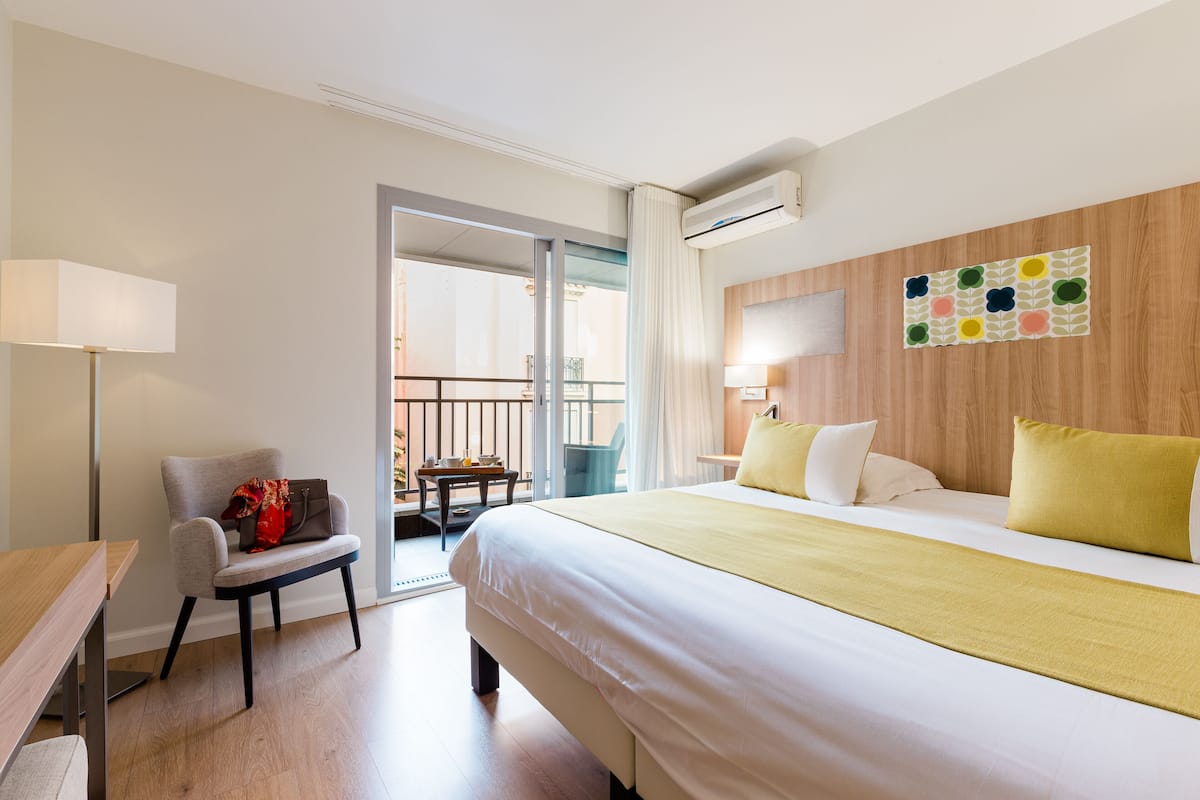 A hotel room with a bed, a chair, a desk, a balcony, and a wall-mounted TV.