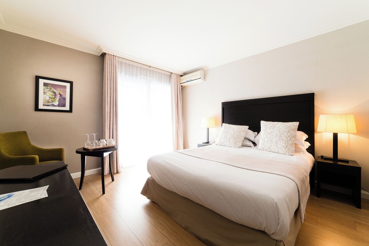 Habitación de hotel con una cama grande, un escritorio con silla, una mesita de noche con lámpara y una ventana con cortinas.