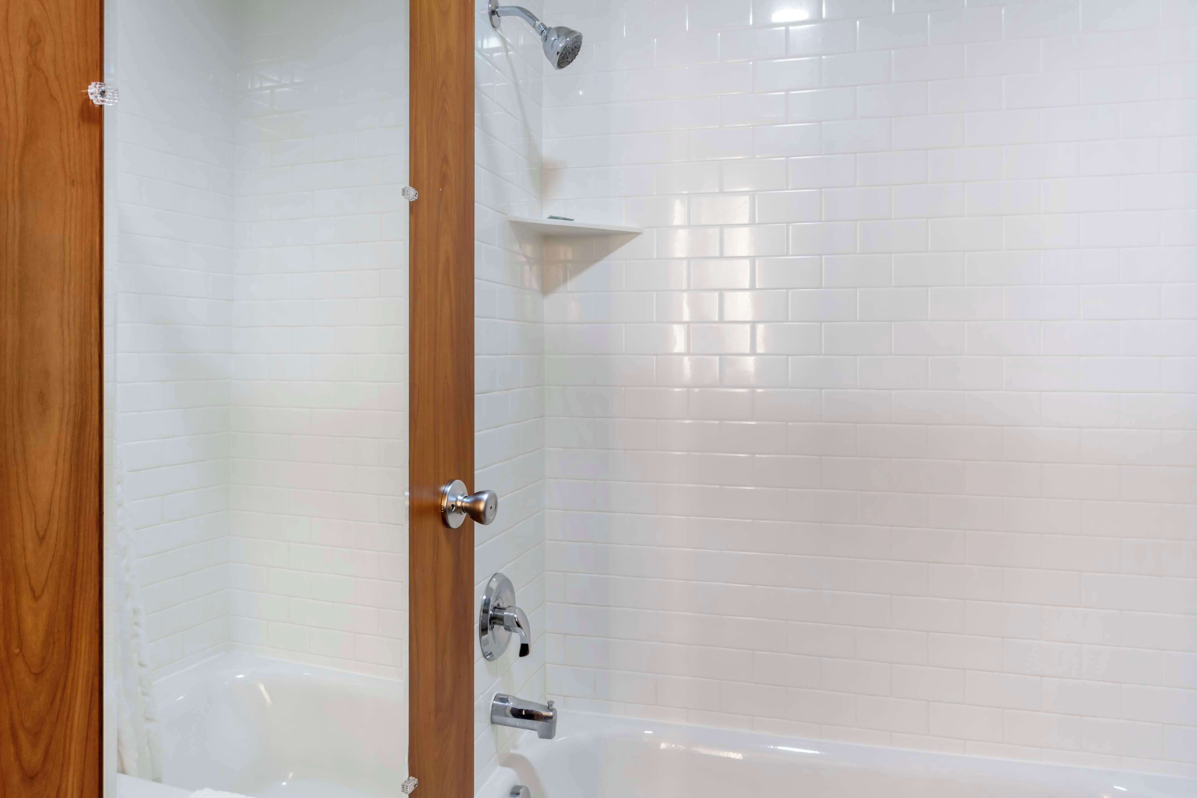 A bathroom with white tiled walls, a wooden door, a bathtub, and a showerhead.