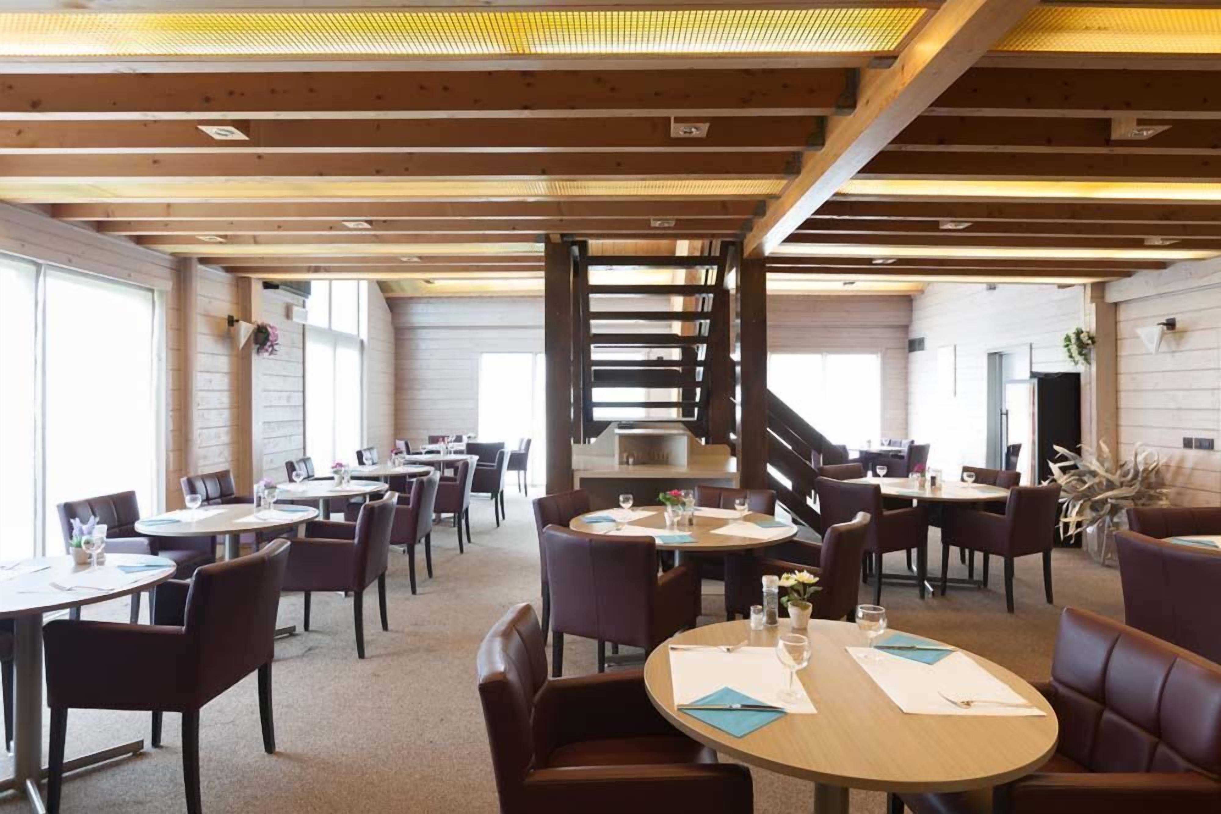 Un comedor espacioso con varias mesas redondas y sillas, vigas de madera en el techo y grandes ventanales que dejan entrar luz natural.