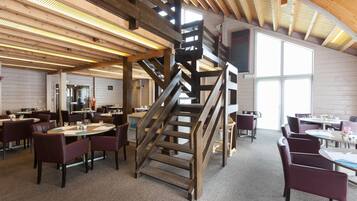 Un vaste espace dédié à la salle à manger, agrémenté d’un escalier central en bois, de tables avec des chaises et de grandes fenêtres qui laissent entrer la lumière naturelle.