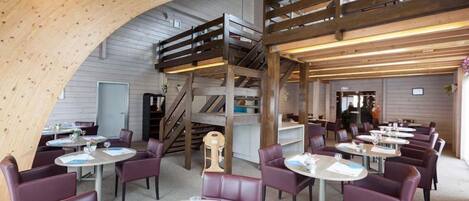 A restaurant with round tables and purple chairs, a wooden staircase, and a high ceiling with exposed beams.