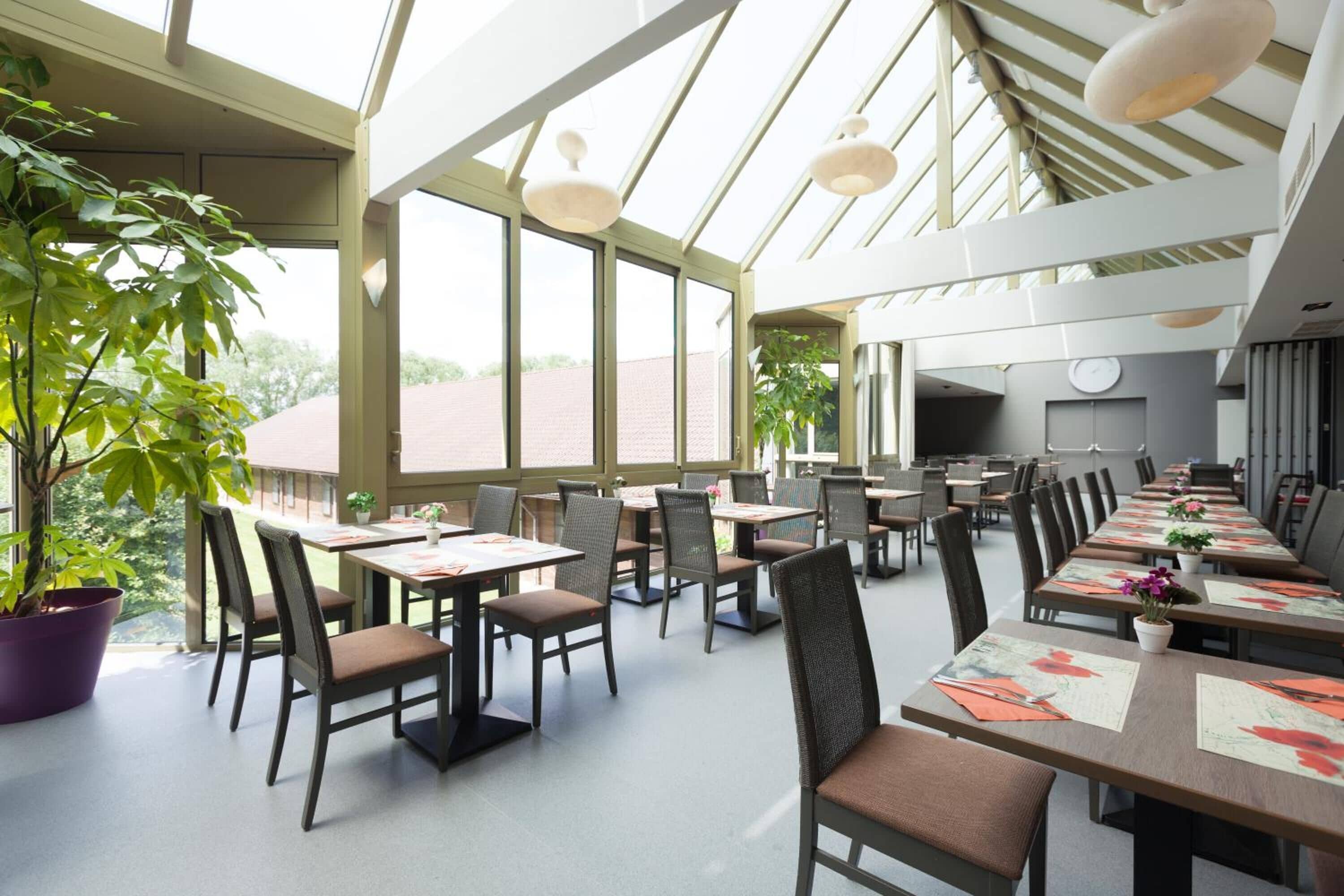 A spacious dining area with long tables, chairs, and potted plants.