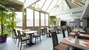 A spacious dining area with long tables, chairs, and potted plants.