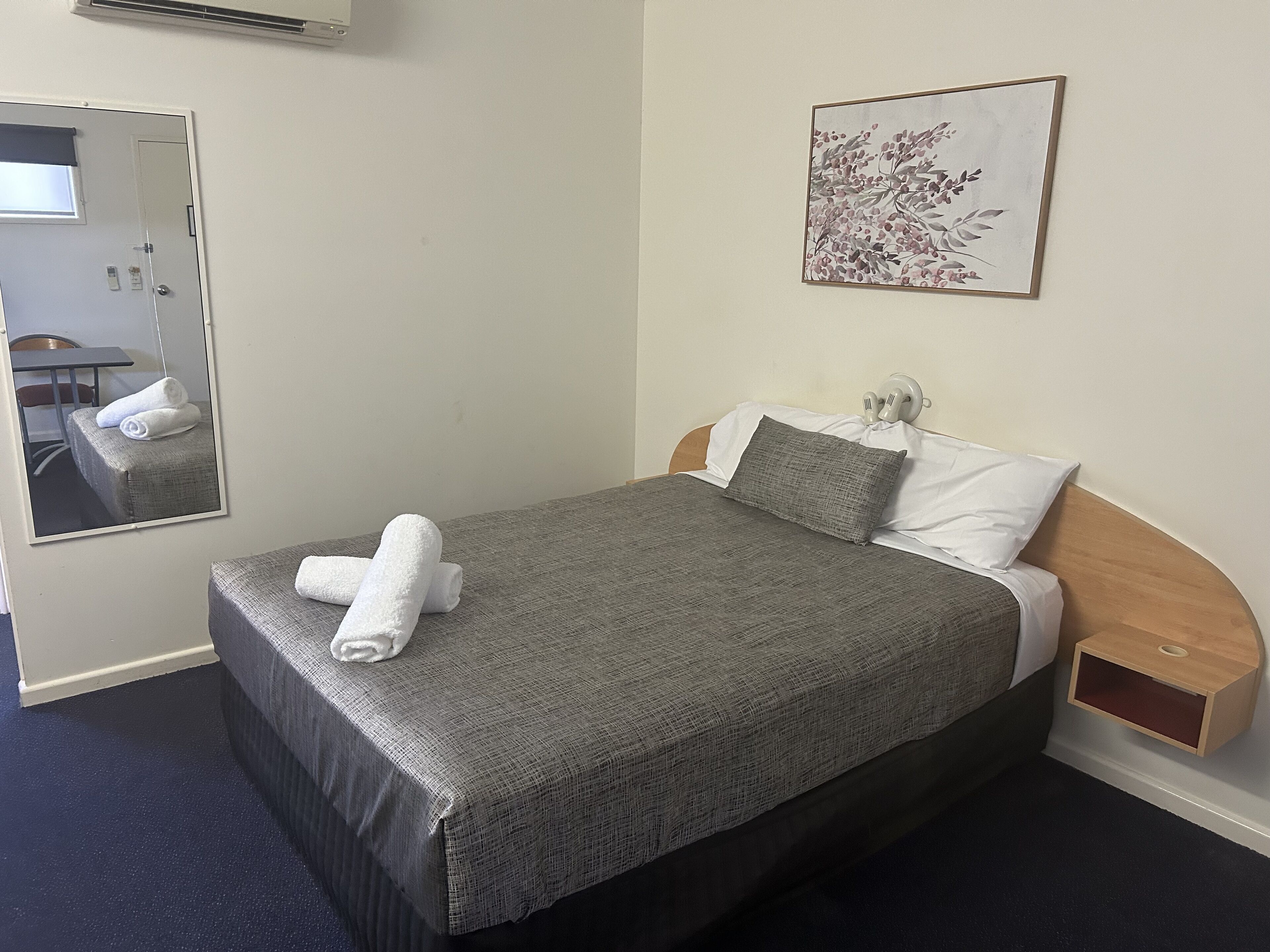 A neatly made bed with a gray blanket and two pillows, a small bedside table, a wall-mounted air conditioner, a framed floral artwork, and a mirror reflecting part of the room.