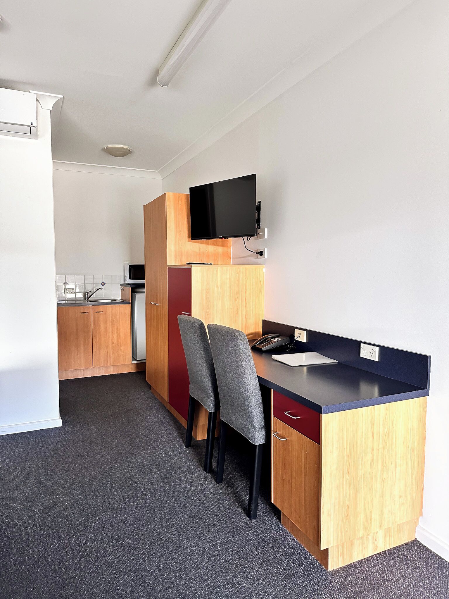 A compact kitchenette with a microwave, a small refrigerator, and a wooden cabinet. There is a desk with two chairs and a flat-screen TV mounted above it.