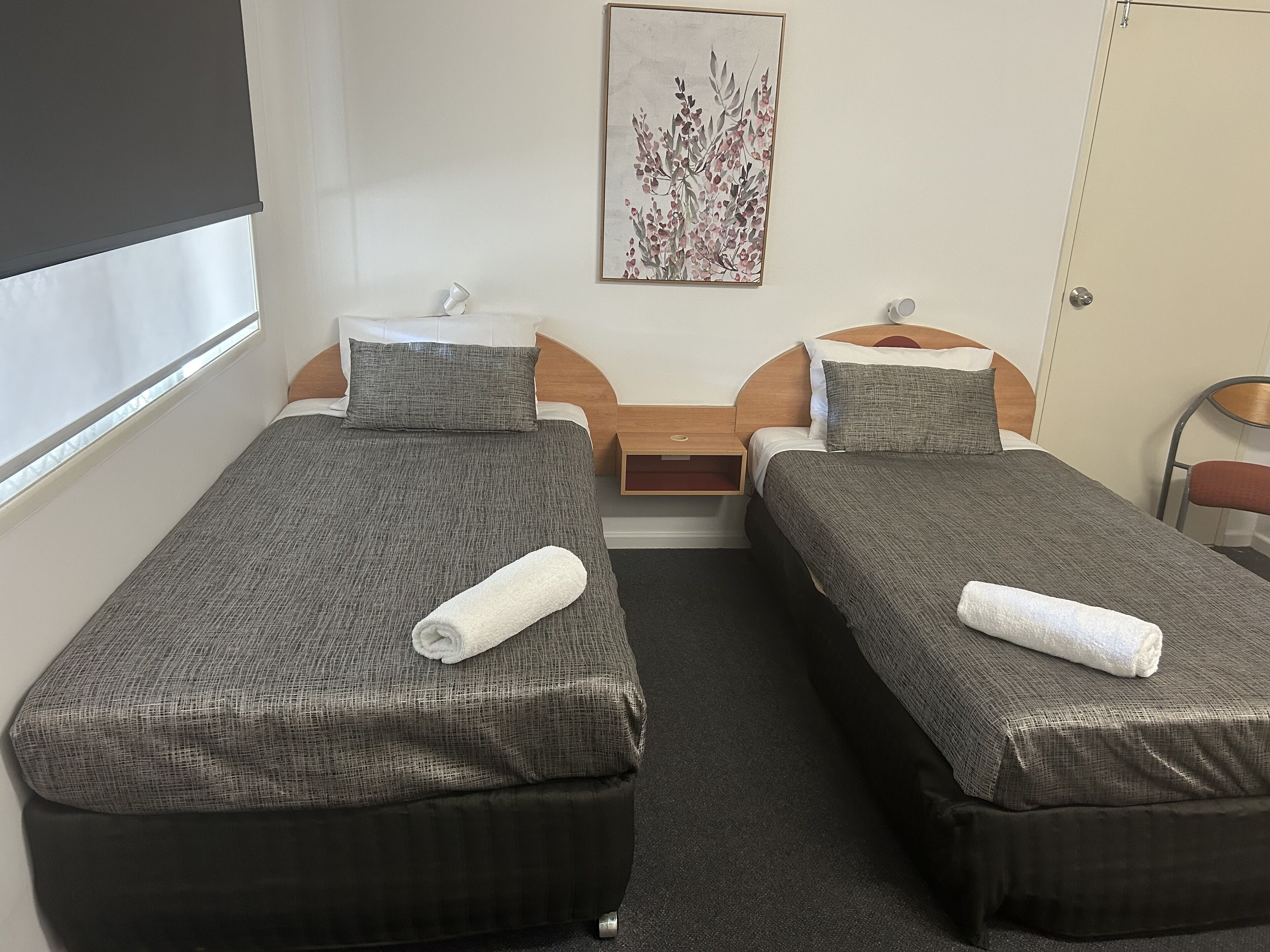 Une chambre double avec deux lits simples, chacun recouvert d’une couverture grise et agrémenté d’une serviette blanche pliée. Une petite table de chevet en bois se trouve entre les lits, une œuvre d’art blanche est accrochée au mur au-dessus de la table de chevet, et une porte se trouve sur le côté droit.